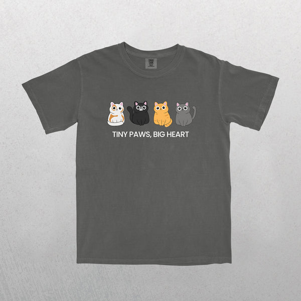 The ‘Tiny Paws, Big Heart’ pet t-shirt in grey features four colorful cartoon cats above the phrase “TINY PAWS, BIG HEART” in white—a cute, funny shirt and perfect gift for pet lovers. Background: light textured surface.