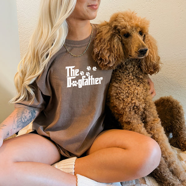 A smiling woman with long blonde hair sits cross-legged holding a brown poodle. She wears The Dogfather shirt with paw prints, cream socks, and has a tattoo—an ideal gift for pet lovers.
