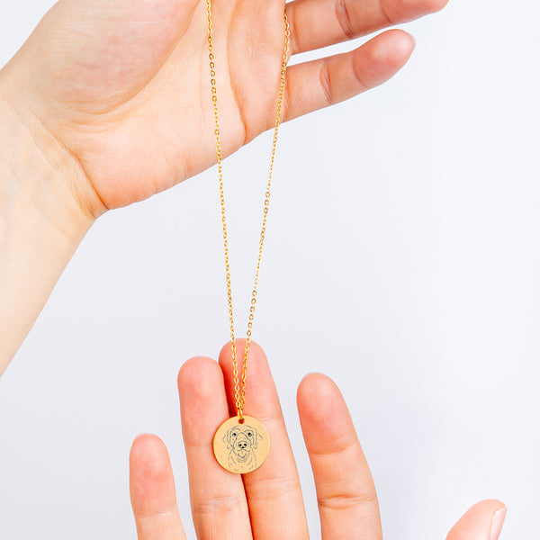 A hand displays the Dog Portrait Necklace - Coin Necklace, featuring a round gold pendant engraved with a dogs face against a white background—an elegant memorial keepsake to honor your beloved pet.