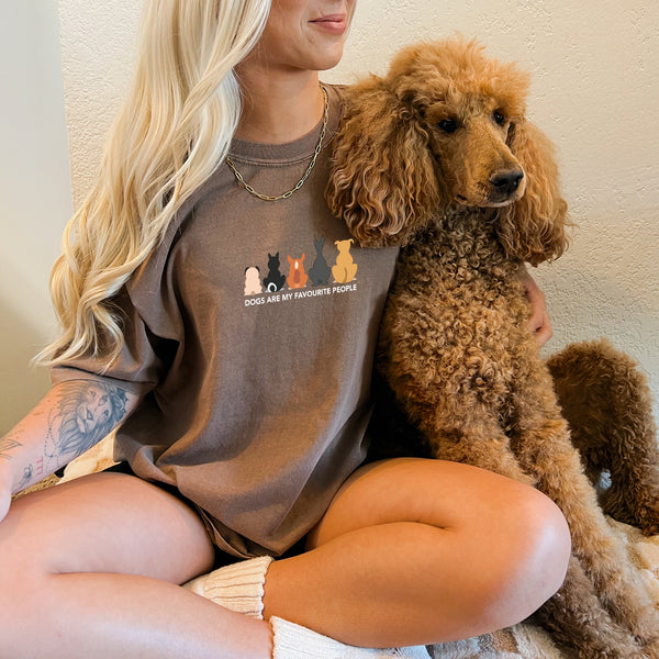 A smiling woman with long blonde hair hugs a brown poodle while wearing the “Dogs are my Favorite People” T-shirt, featuring cartoon dog illustrations—a fun pet tee for any animal lover.