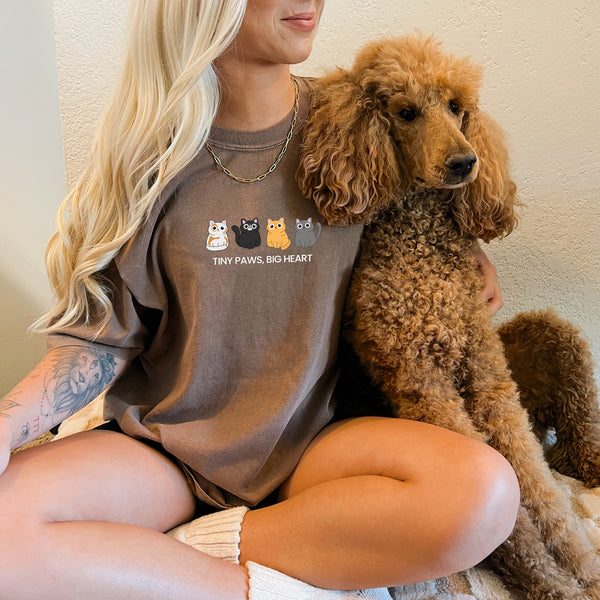 A smiling, tattooed woman with long blond hair sits cross-legged, wearing the Tiny Paws, Big Heart shirt with cartoon animals. She hugs a curly-haired brown poodle—an ideal gift for any pet lover.