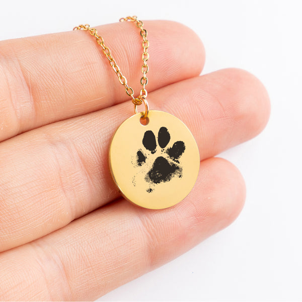 A hand holds the Dog Paw Necklace - Coin Necklace, featuring a round gold pendant with a black paw print—making it a perfect personalized pet jewelry piece and thoughtful gift for pet lovers, displayed on a plain white background.