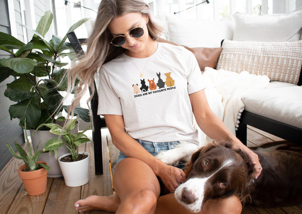 Relaxing on a cozy porch, a woman wears the Dogs are my Favorite People vintage pet t-shirt and pets her brown and white dog. Surrounded by plants and cushions, its a charming scene—an ideal gift for any pet lover.