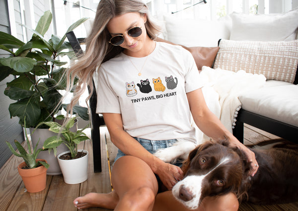 A woman in sunglasses and a Tiny Paws, Big Heart vintage pet t-shirt sits on a sofa, petting her brown and white dog. Green potted plants decorate the porch, creating a charming scene with this unique pet keepsake.