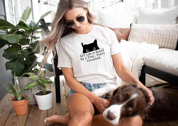 A woman in sunglasses and a Tiny Furry Overlord t-shirt sits on the porch petting her brown and white dog, with a potted plant and beige sofa nearby—an ideal scene for showcasing this fun pet lover tee.