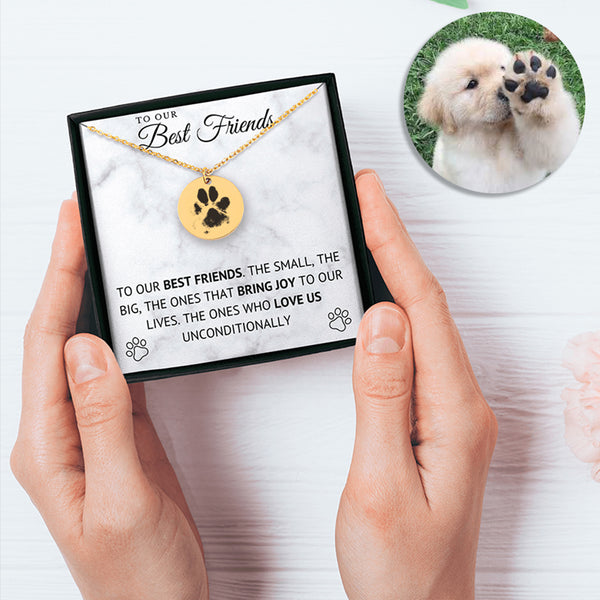 A person holds a Dog Paw Necklace - Coin Necklace, a personalized pet jewelry piece in a box that says “To our best friends...” with a heartfelt message. An inset photo features a fluffy puppy raising its paw.