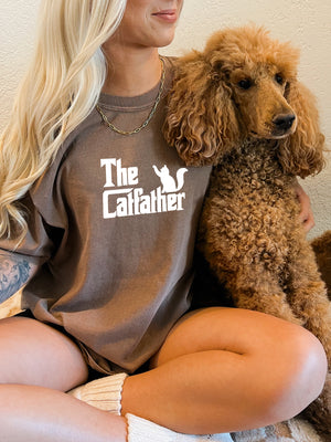 A tattooed woman with long blonde hair sits cross-legged, cuddling a large brown poodle while wearing The Catfather t-shirt—a unique keepsake for cat lovers.