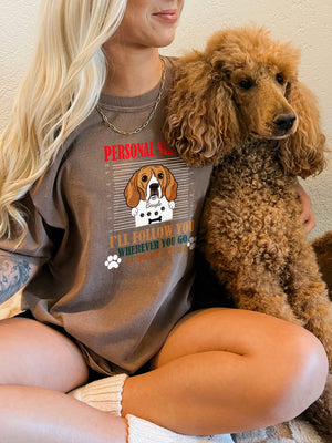 A smiling blonde woman sits cross-legged holding a brown poodle, wearing the Personal Stalker shirt featuring the phrase “I’ll Follow You Wherever You Go” and a dog illustration.