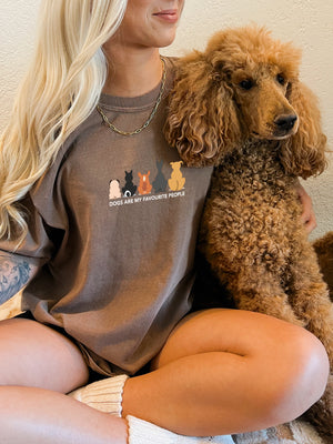 A smiling woman with long blonde hair hugs a brown poodle while wearing the “Dogs are my Favorite People” T-shirt, featuring cartoon dog illustrations—a fun pet tee for any animal lover.