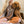 A smiling, tattooed woman with long blond hair sits cross-legged, wearing the Tiny Paws, Big Heart shirt with cartoon animals. She hugs a curly-haired brown poodle—an ideal gift for any pet lover.