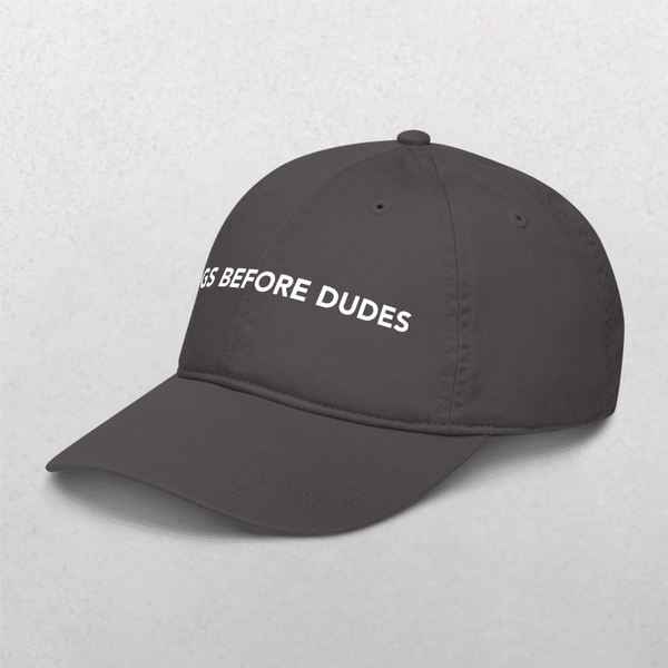 The Classic Organic Dad Cap - Dogs Before Dudes features dark gray fabric with white DOGS BEFORE DUDES embroidery, making it a perfect slogan pet hat for any dog mom. Displayed against a light gray background.