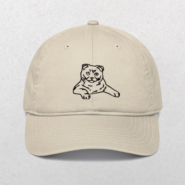 The Embroidered Line art Pet Portrait Organic Classic Dad Cap in beige features a simple black line drawing of a lounging cat embroidered on the front, set against a light, textured background.
