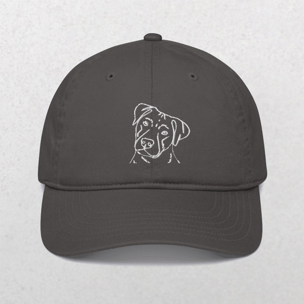 The Embroidered Line Art Pet Portrait Organic Classic Dad Cap is a dark gray hat with a white embroidered dog face on the front center, shown on a light textured background.