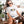 A woman in sunglasses and a Tiny Paws, Big Heart vintage pet t-shirt sits on a sofa, petting her brown and white dog. Green potted plants decorate the porch, creating a charming scene with this unique pet keepsake.