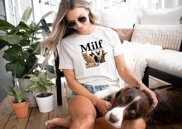 A woman in sunglasses sits on a couch petting a brown and white dog. She wears the Milf vintage pet t-shirt, featuring three cats below the text—a perfect gift for any pet lover. Green plants and cozy decor complete the scene.