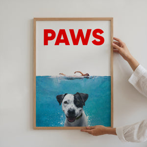 A person hangs the Paws Film Cover canvas—a playful artwork mimicking the Jaws poster, featuring a swimmer above and dog below with PAWS in bold red. This fun pet decor is a perfect gift for dog lovers.
