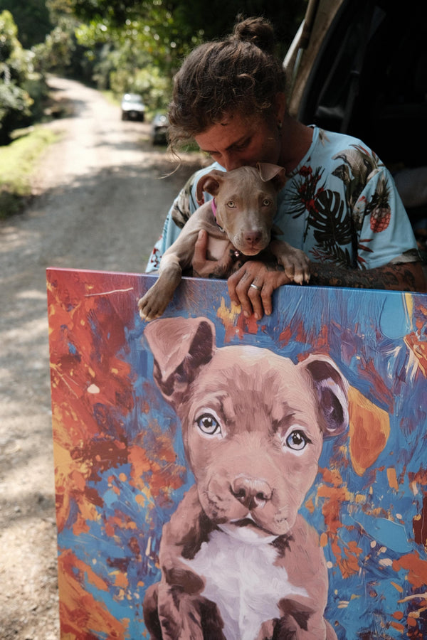 A person holds a puppy and an Oil Masterpiece - Custom Pet Portrait of the same puppy, standing by a dirt road with trees. Looking at the puppy affectionately, they showcase this perfect gift for any pet lover.