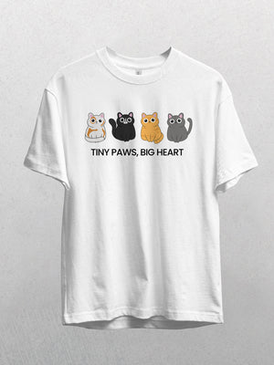 The white Tiny Paws, Big Heart shirt displays four colorful cartoon cats above the slogan, perfect for cat lovers. Shown hanging against a light gray background.