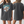 A woman models the Personalised Retro Tee in blue. The loose-fitting shirt features a front graphic of a piglet with clouds and WILBUR above it; the back is plain. She has long, light brown hair and wears bracelets.