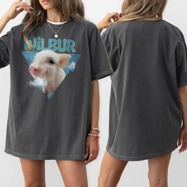 A woman models the Personalised Retro Tee in blue. The loose-fitting shirt features a front graphic of a piglet with clouds and WILBUR above it; the back is plain. She has long, light brown hair and wears bracelets.